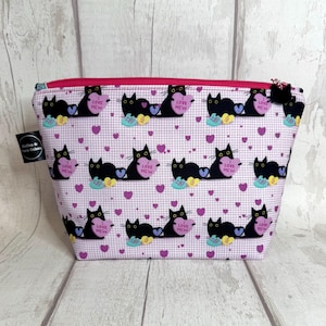 May include: Pink zippered makeup bag with a black cat print. The cats are holding pink hearts with the text "I Love Mew" on them. The background is a white and pink checkered pattern with small pink hearts.