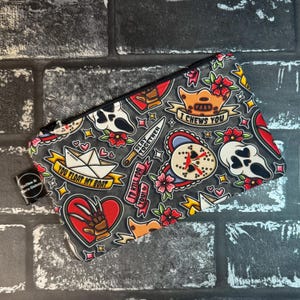May include: Black and white zippered pouch with a horror movie themed print. The print features hearts, knives, and characters from horror movies. The text on the pouch reads "You float my boat", "I'd kill for you", "Slay together", and "I chews you".
