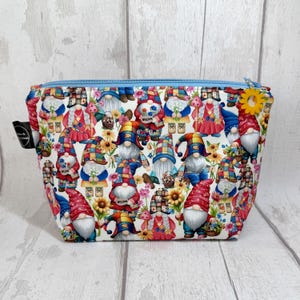 Può includere: Un beauty case con cerniera con un motivo stravagante di gnomi, girasoli ed elementi floreali su sfondo bianco. La borsa ha una cerniera blu chiaro e un tirazip a forma di fiore giallo. Per riporre trucchi e prodotti di bellezza.