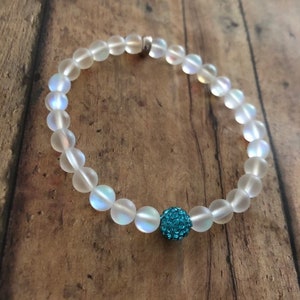 May include: A beaded bracelet featuring iridescent, frosted glass beads. A single, round, turquoise-colored bead with a textured surface is centered on the bracelet. The bracelet is displayed on a wooden surface.