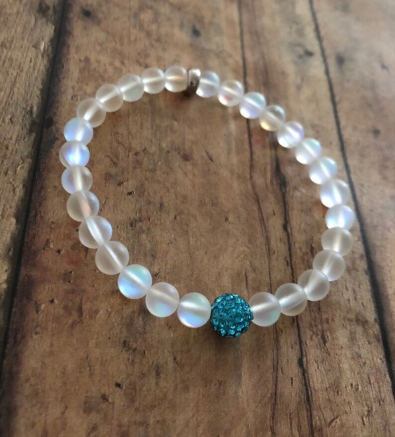 May include: A beaded bracelet featuring iridescent, frosted glass beads. A single, round, turquoise-colored bead with a textured surface is centered on the bracelet. The bracelet is displayed on a wooden surface.