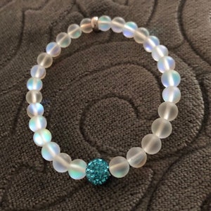 May include: A clear beaded bracelet with a single, round, turquoise-colored bead. The bracelet is made of translucent, round beads with a subtle iridescent sheen. The clasp is silver-colored.