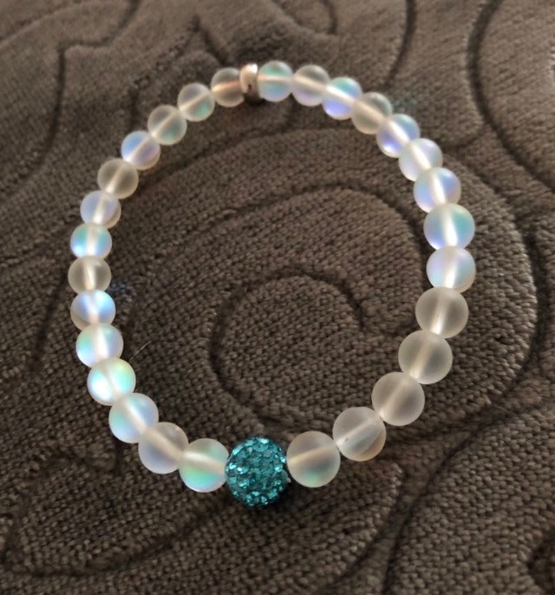 May include: A clear beaded bracelet with a single, round, turquoise-colored bead. The bracelet is made of translucent, round beads with a subtle iridescent sheen. The clasp is silver-colored.