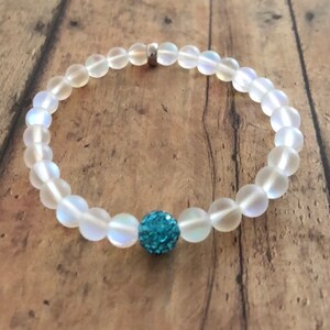 May include: A beaded bracelet featuring iridescent, round beads with a central turquoise, sparkly accent bead. The bracelet is strung on a clear elastic cord and has a silver-colored clasp.