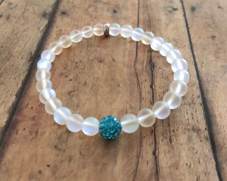 May include: A beaded bracelet featuring iridescent, round beads with a central turquoise, sparkly accent bead. The bracelet is strung on a clear elastic cord and has a silver-colored clasp.