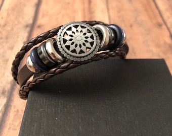 May include: Braided brown leather bracelet with a silver-tone centerpiece featuring a floral design. The bracelet includes silver and navy blue accents. The bracelet is displayed on a black surface.