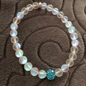 May include: A beaded bracelet featuring iridescent, frosted glass beads. The bracelet includes a single, larger, round bead covered in small, sparkling, light blue crystals. The bracelet is strung on a clear elastic cord and has a silver clasp.