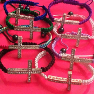 May include: A collection of adjustable cord bracelets featuring silver-toned cross charms encrusted with clear rhinestones. The woven bands come in various colors including blue, green, purple, pink, white, and brown, each accented with small dark beads.