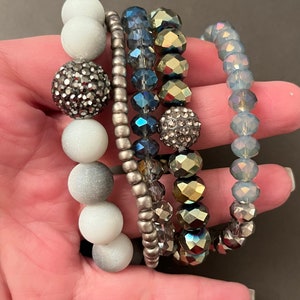 May include: A set of four stretch bracelets featuring a variety of beads. One strand has matte white and glittery dark gray spheres. Another has small metallic silver beads. The third has faceted blue and gold beads. The fourth has light blue crystals.