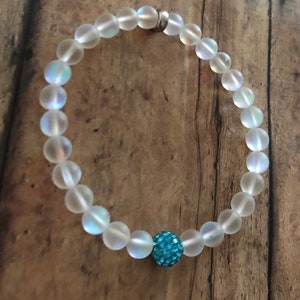 May include: A beaded bracelet featuring iridescent, frosted glass beads. The bracelet has a single, round, turquoise-colored, sparkly bead. The bracelet is strung on a clear elastic cord and is displayed on a wooden surface.