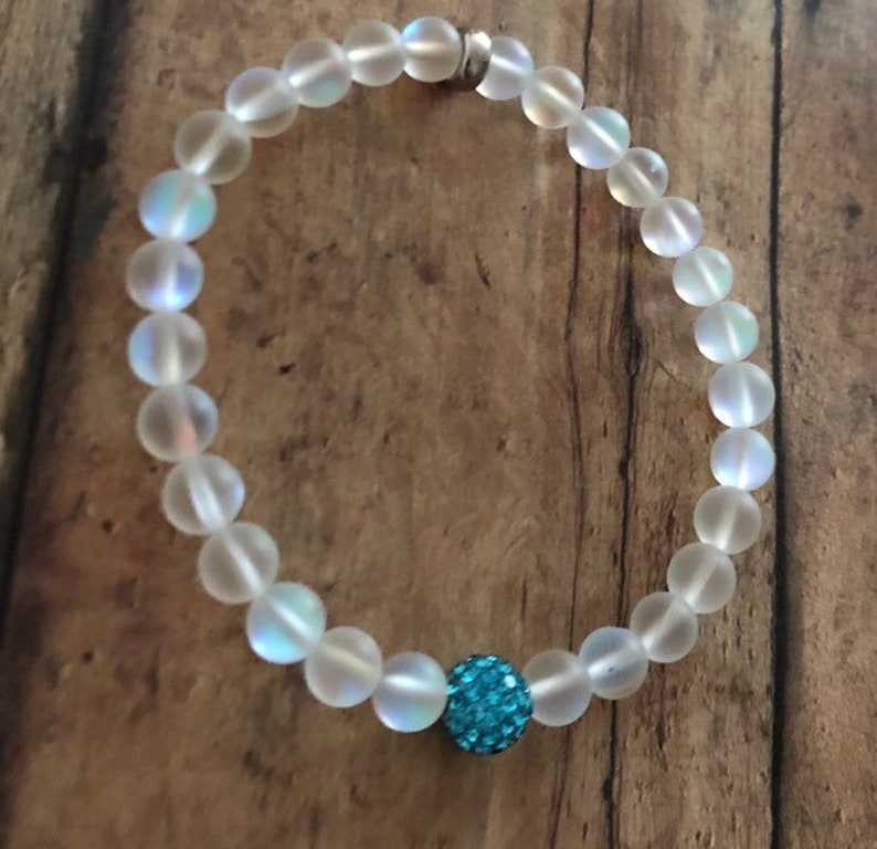 May include: A beaded bracelet featuring iridescent, frosted glass beads. The bracelet has a single, round, turquoise-colored, sparkly bead. The bracelet is strung on a clear elastic cord and is displayed on a wooden surface.