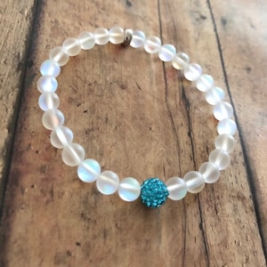 May include: A clear, iridescent beaded bracelet with a silver clasp. The bracelet features round, translucent beads and a single, turquoise-colored, sparkly accent bead. The bracelet is displayed on a wooden surface.