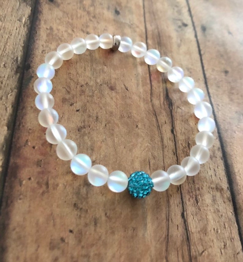 May include: A clear, iridescent beaded bracelet with a silver clasp. The bracelet features round, translucent beads and a single, turquoise-colored, sparkly accent bead. The bracelet is displayed on a wooden surface.