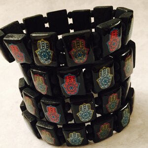 May include: A black elastic bracelet with multiple rows of colorful hamsa hand charms. Each charm features a blue eye in the center.