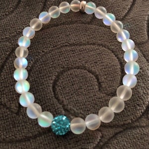 May include: A beaded bracelet featuring iridescent, frosted glass beads. The bracelet includes a single, larger, sparkly blue bead. A small silver-colored metal spacer is also present. The bracelet is displayed on a textured, gray surface.