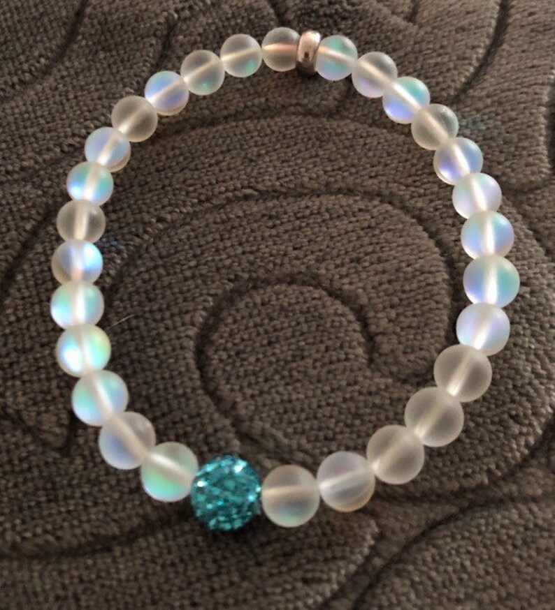 May include: A beaded bracelet featuring iridescent, frosted glass beads. The bracelet includes a single, larger, sparkly blue bead. A small silver-colored metal spacer is also present. The bracelet is displayed on a textured, gray surface.