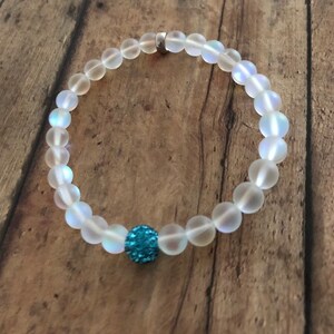 May include: A beaded bracelet featuring translucent, round beads with a central blue, sparkly accent bead. The bracelet is strung on a clear elastic band and has a silver clasp. The bracelet is displayed on a wooden surface.