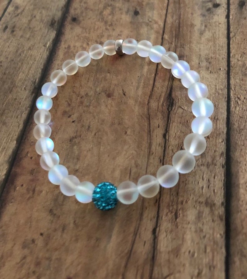 May include: A beaded bracelet featuring translucent, round beads with a central blue, sparkly accent bead. The bracelet is strung on a clear elastic band and has a silver clasp. The bracelet is displayed on a wooden surface.