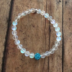 May include: A beaded bracelet featuring iridescent, frosted glass beads. The bracelet has a single, round, turquoise-colored bead with a sparkly, textured surface. The bracelet is strung on a clear elastic cord and has a silver-colored clasp.