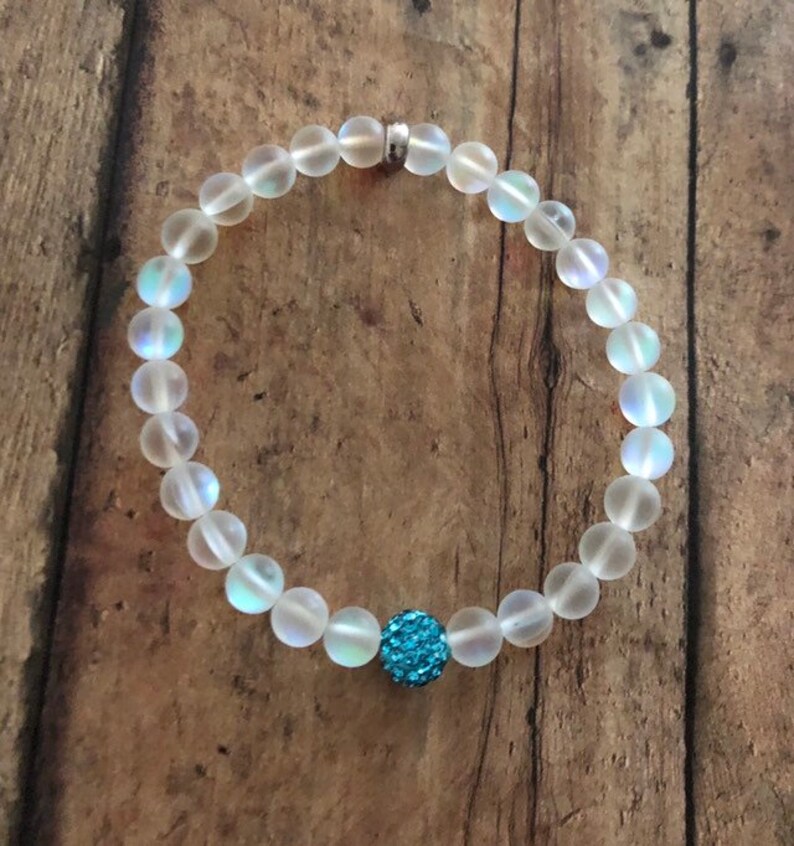 May include: A beaded bracelet featuring iridescent, frosted glass beads. The bracelet has a single, round, turquoise-colored bead with a sparkly, textured surface. The bracelet is strung on a clear elastic cord and has a silver-colored clasp.