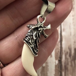 May include: A pendant featuring a curved, off-white tooth-shaped element set into a silver-toned metal cap. The metal cap is sculpted with intricate, raised detailing and is attached to a black cord by a polished silver-toned metal bail.
