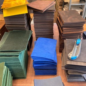 May include: Stacks of different coloured leather sheets, including green, blue, brown, and yellow. The leather sheets are arranged on a wooden surface. A utility knife is lying on top of one of the stacks.