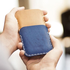 Può includere: Una custodia per telefono in pelle blu con una patta in pelle marrone. La custodia ha cuciture bianche sui bordi.