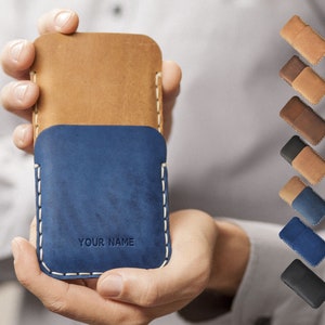 Può includere: Una custodia per telefono in pelle blu con una patta in pelle marrone. La custodia ha cuciture bianche e il testo "YOUR NAME" è in rilievo sul davanti. La custodia è tenuta nella mano di una persona.