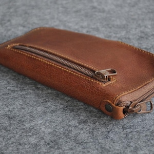 Könnte beinhalten: Eine braune Leder-Reißverschlusstasche mit einem einzigen Reißverschluss-Zipper. Die Tasche liegt auf einer grauen, strukturierten Oberfläche.