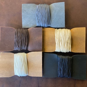 Op de afbeelding: Zes spoelen met gewaxt garen, gewikkeld in leer. De spoelen zijn in een piramidevorm gerangschikt. De garenkleuren zijn grijs, bruin, beige, wit, zwart en donkergrijs.