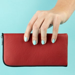 May include: A red leather wallet with a zipper closure. The wallet is being held by a hand with light blue nail polish.