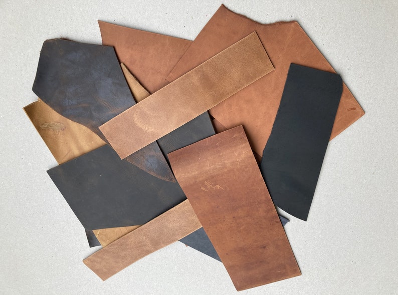 May include: Assortment of leather scraps in various shades of brown and black. The leather pieces are arranged in a pile on a white surface.
