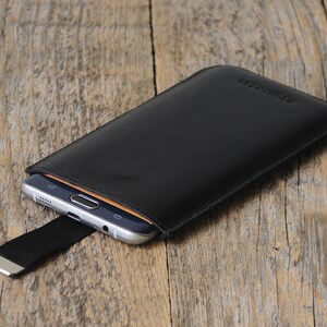 May include: Black leather phone case with a stitched edge and a black elastic strap with a silver metal clasp. The case has the word "Genuine Leather" embossed on the front.