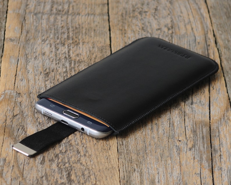 May include: Black leather phone case with a stitched edge and a black elastic strap with a silver metal clasp. The case has the word "Genuine Leather" embossed on the front.