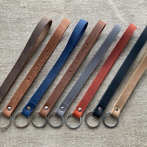 Peut inclure: Plusieurs porte-clés en cuir de différentes couleurs, dont marron, bleu, gris et rouge. Certains porte-clés portent l'inscription "YOUR TEXT HERE". Chaque porte-clés est équipé d'un anneau métallique.