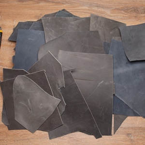 May include: A pile of dark gray and black leather scraps on a wooden surface. A yellow utility knife is visible to the left of the pile.