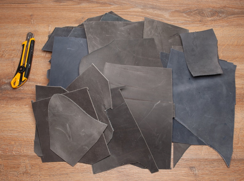 May include: A pile of dark gray and black leather scraps on a wooden surface. A yellow utility knife is visible to the left of the pile.