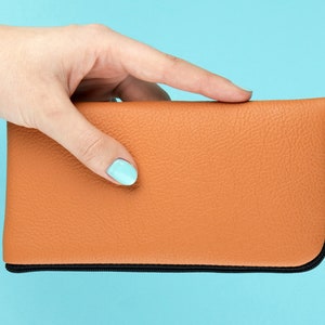 Può includere: Una pochette in pelle marrone con cerniera nera. La pochette è tenuta da una mano con smalto per unghie blu chiaro.
