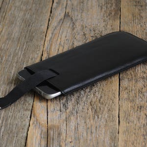 May include: A black leather phone case with a black strap and a silver metal clasp. The case is lying on a wooden surface.