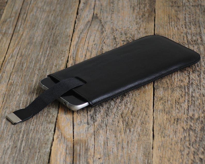 May include: A black leather phone case with a black strap and a silver metal clasp. The case is lying on a wooden surface.