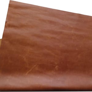 Puede incluir: Una pieza rectangular de cuero marrón medio con un acabado liso y mate en la parte superior y una parte inferior de ante texturizado y fibroso visible en la esquina. La superficie muestra variaciones naturales de color y marcas de grano.