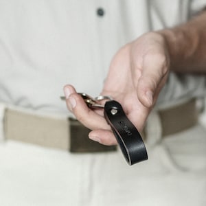 Könnte beinhalten: Ein schwarzer Leder-Schlüsselanhänger mit dem eingeprägten Wort "BRAVO". Der Schlüsselanhänger ist an einem Schlüsselsatz befestigt.