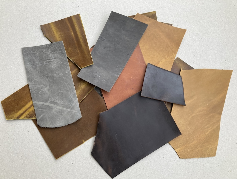 May include: Assortment of leather scraps in various colors and textures, including brown, gray, black, and tan. The leather pieces are arranged in a pile on a white surface.