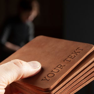 May include: A stack of square, brown leather coasters. The top coaster has the words "YOUR TEXT" engraved on it. The coasters are held in a hand, with a blurred background.