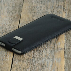 May include: Black leather phone case with a zipper closure and a silver metal clasp. The case is lying on a wooden surface.