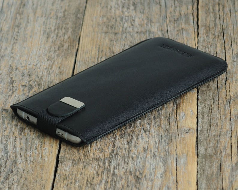 May include: Black leather phone case with a zipper closure and a silver metal clasp. The case is lying on a wooden surface.