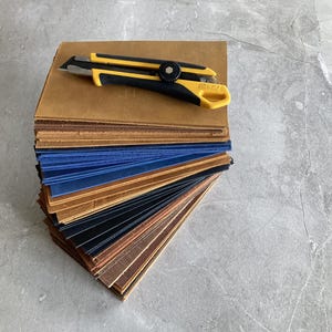 Könnte beinhalten: Ein Stapel Lederproben in verschiedenen Farben, darunter Braun, Blau und Schwarz, mit einem gelb-schwarzen Teppichmesser darauf. Die Lederproben sind fächerförmig angeordnet und zeigen die verschiedenen Texturen und Farbtöne.