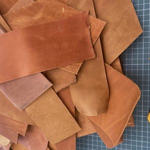 May include: A pile of brown leather scraps of various sizes and shapes. A yellow utility knife is lying on a green cutting mat.