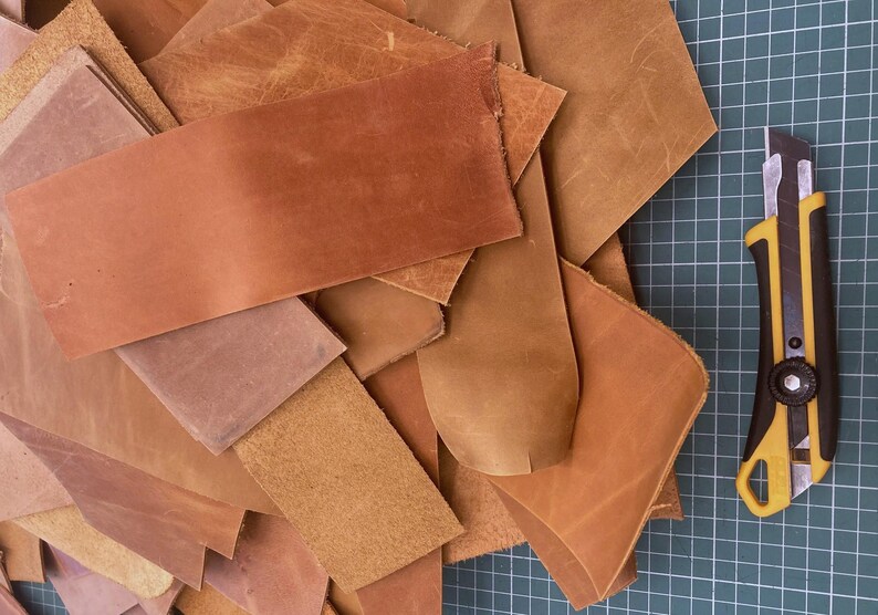 May include: A pile of brown leather scraps of various sizes and shapes. A yellow utility knife is lying on a green cutting mat.