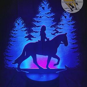 May include: A blue and pink LED night light with a silhouette of a person riding a horse in front of a forest. The light is on a round base with a switch. The text "www.yellaponycustoms.com" is at the bottom of the image.